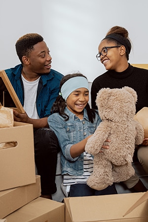 Young Afro Caribbean man and woman smiling at each other surrounded by brown packing boxes sitting with young girl in denim jacket looking at large teddy bear just lifted from packing box