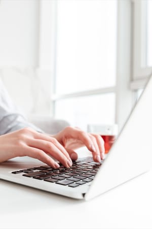 Close up of young woman's hands typing on a laptop on a white desk with window and glass of herbal tea in the background