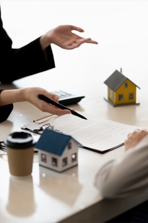 Close up of business woman's upturned hands one holding a pen opposite man's arm in shirt sleeve with clipboard paperwork on a desk two model houses calculator and take away coffee cup