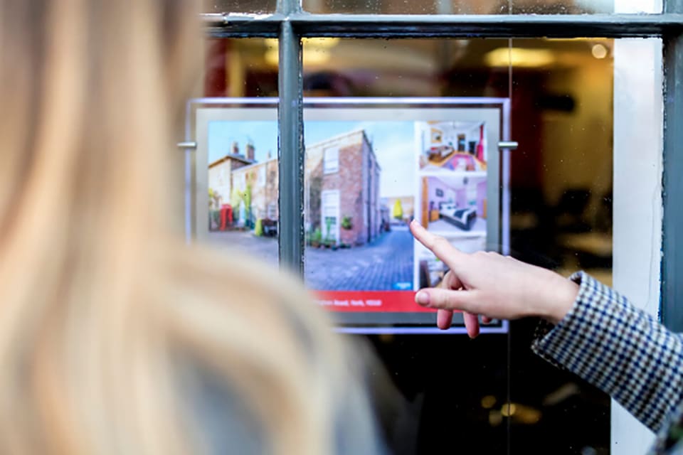 Close up of the back of the head of a blonde long haired person and a young persons hand pointing to property details in a shop window