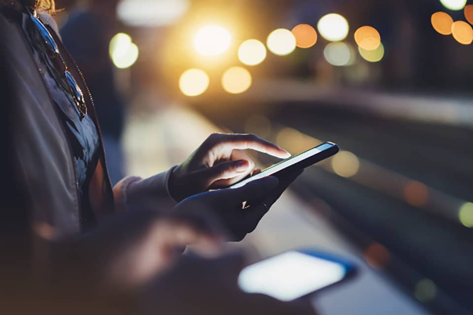 Evening image of woman touching backlit mobile phone screen with dappled street lights in the background