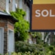 Close up of brown and yellow sold sign in front of traditional brick terraced home with bay window and lush green frontages