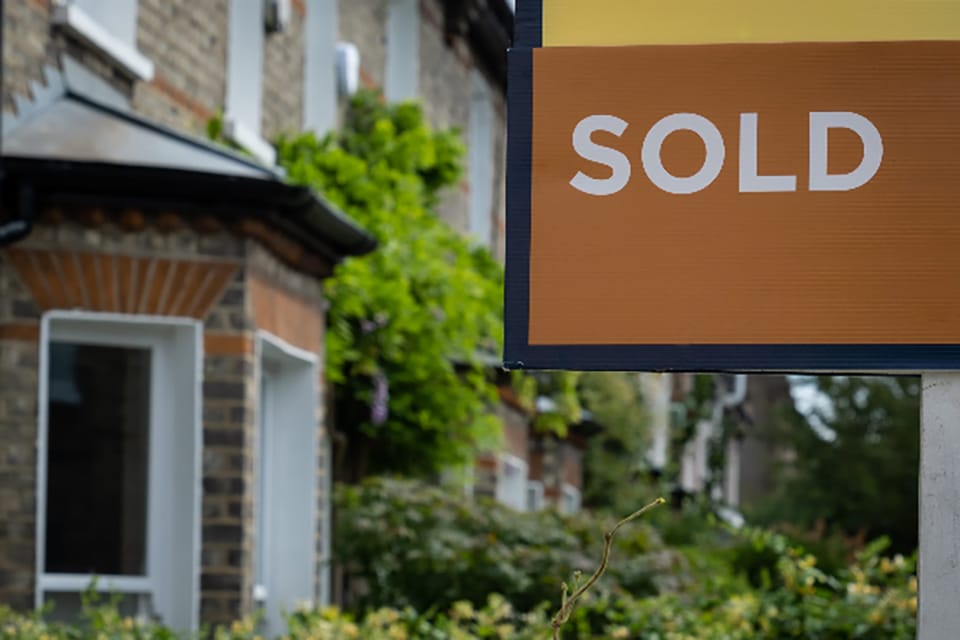 Close up of brown and yellow sold sign in front of traditional brick terraced home with bay window and lush green frontages