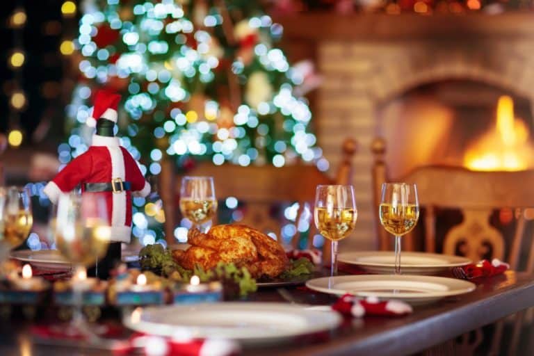 Christmas styled dining table set with plates glasses of white wine tealights Santa decorations and roast chicken with lit Christmas tree and open lit fire in brick fireplace in the background