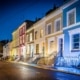 Evening street view image of a row of traditional London town houses painted different colours with stone steps and wrought iron railings decorated with lit fairy lights and Christmas wreath on front door of the nearest home