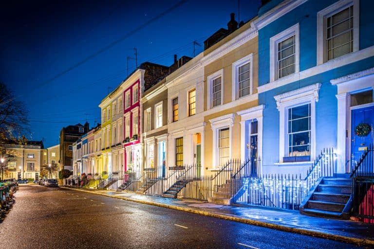 Evening street view image of a row of traditional London town houses painted different colours with stone steps and wrought iron railings decorated with lit fairy lights and Christmas wreath on front door of the nearest home