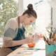 Young woman glazing ceramic pot sitting at a work bench in studio setting with pot of artist brushes in the foreground and large window in the background