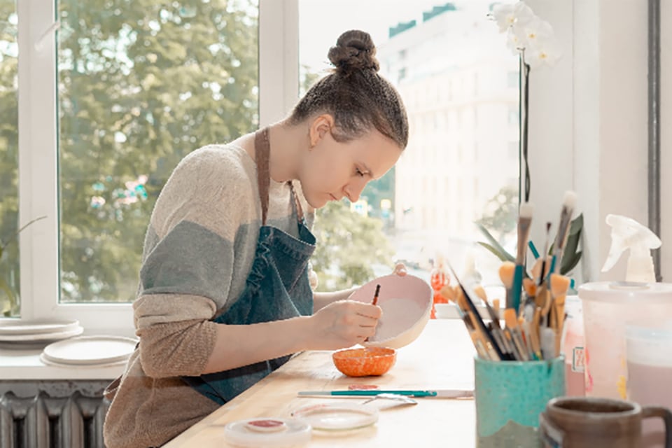 Young woman glazing ceramic pot sitting at a work bench in studio setting with pot of artist brushes in the foreground and large window in the background