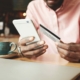 Young Afro Caribbean man sitting at cafe table with closed laptop and artisanal cup and saucer with bank card in one hand and mobile phone in the other