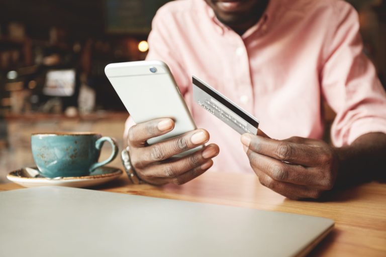 Young Afro Caribbean man sitting at cafe table with closed laptop and artisanal cup and saucer with bank card in one hand and mobile phone in the other