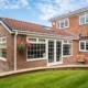 Rear of modern build red brick property with large garden room patio and wooden bench with walled curved lawn hanging baskets planters and flower border