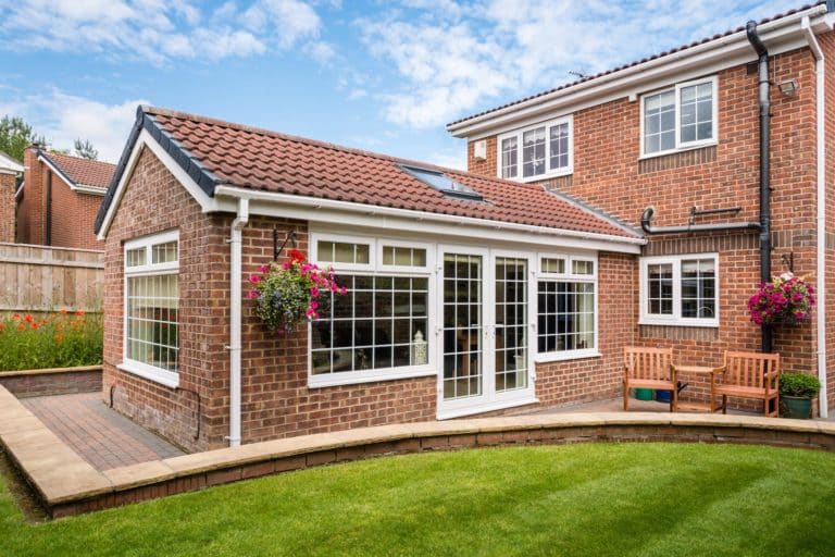 Rear of modern build red brick property with large garden room patio and wooden bench with walled curved lawn hanging baskets planters and flower border