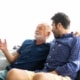 Mature father and son image sitting on sofa with soft lit window behind arms around one another talking and smiling