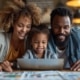 Afro Caribbean bearded man and woman with young child at kitchen table smiling whilst looking at laptop with colourful print outs on the table in the foreground