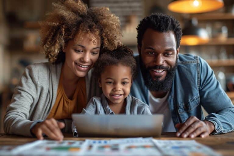 Afro Caribbean bearded man and woman with young child at kitchen table smiling whilst looking at laptop with colourful print outs on the table in the foreground