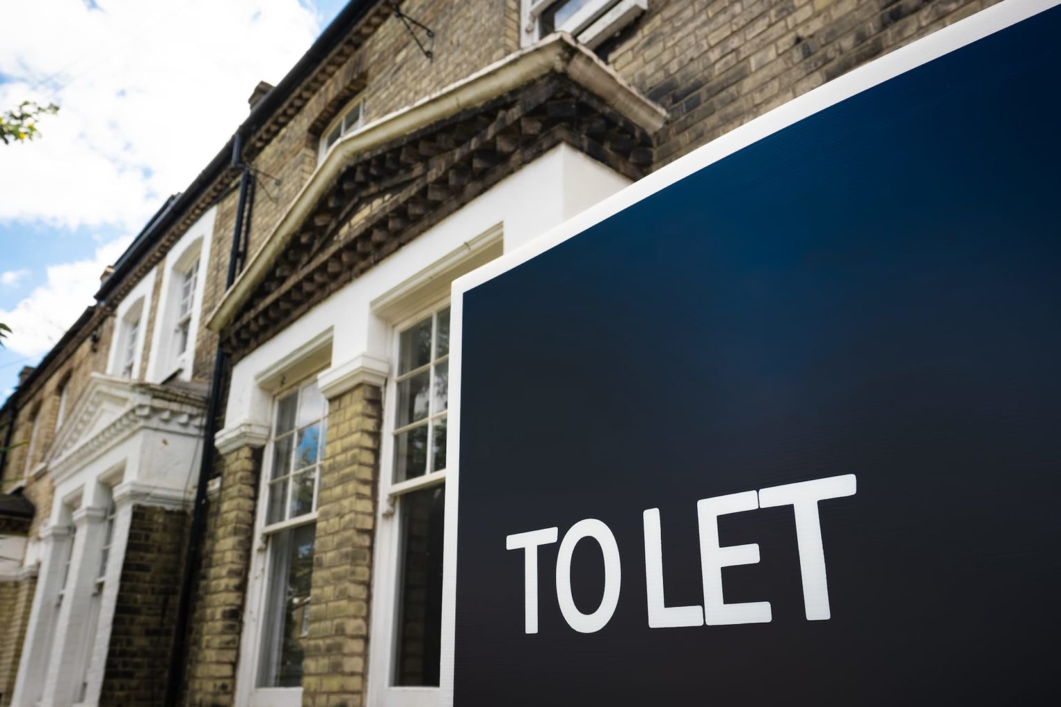 Dark blue To Let sign with white text outside entrance to a period property with stone mullion windows and ornate stone brick work