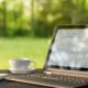 Outdoor table with open laptop white cup and saucer open notepad and pen with green lawn and trees in the background