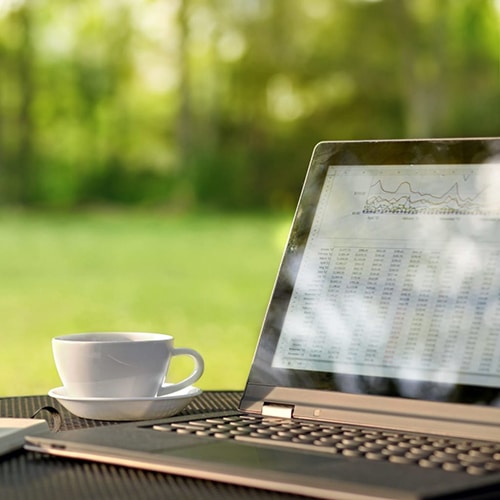 Outdoor table with open laptop white cup and saucer open notepad and pen with green lawn and trees in the background