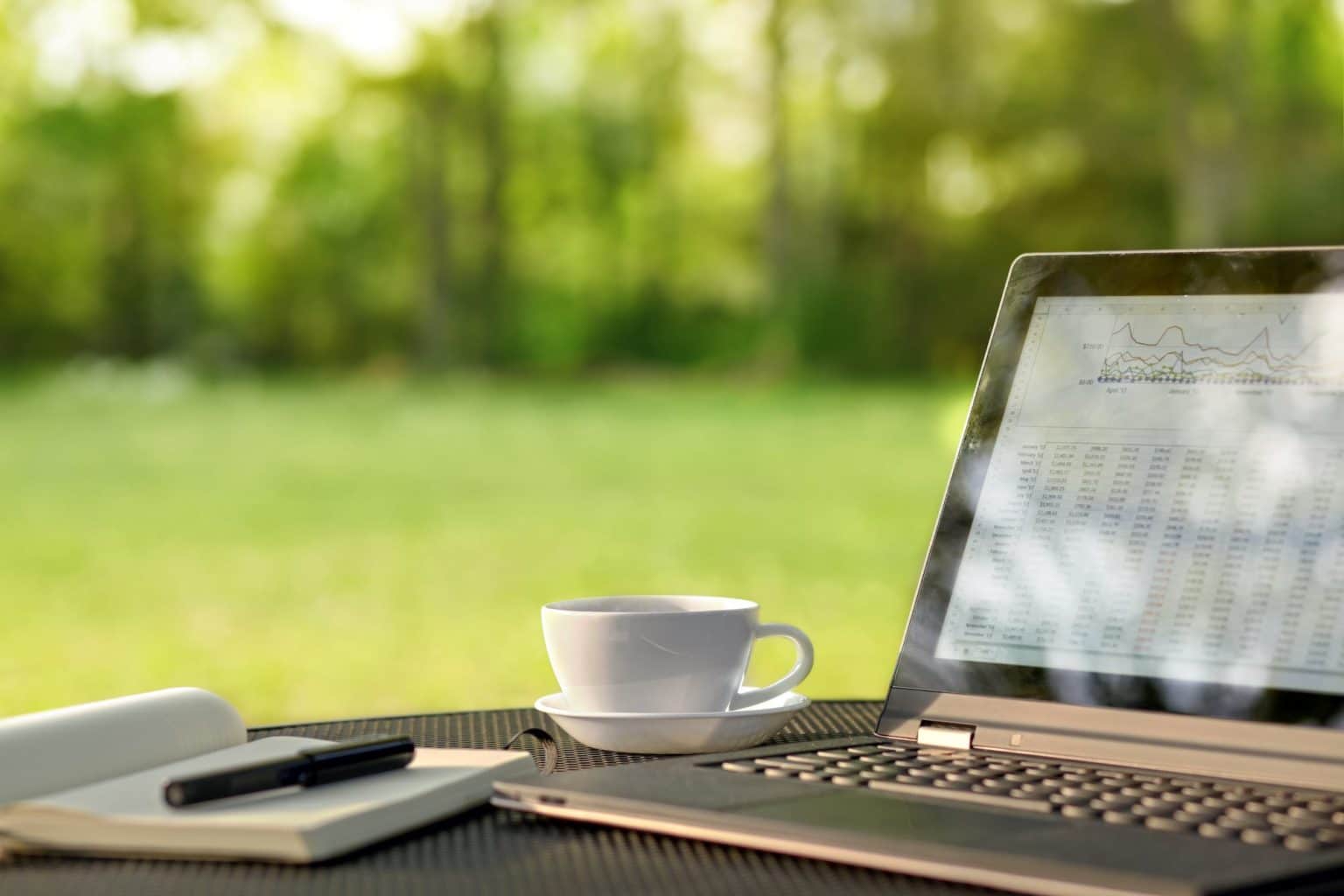 Outdoor table with open laptop white cup and saucer open notepad and pen with green lawn and trees in the background