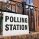 Polling station white sign with black lettering tied to black railings outside entrance to brick community building