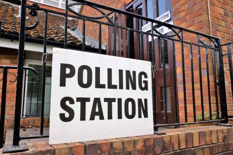 Polling station white sign with black lettering tied to black railings outside entrance to brick community building