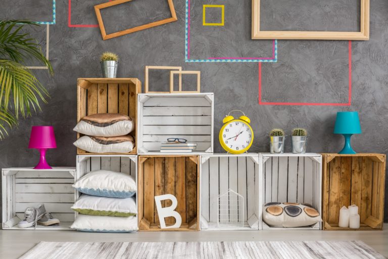 Display of wooden crates some painted white filled with cushions trainers books candles lamps cactus plants letter B frames graphics on wall and stripe textured neutral rug on floor