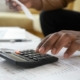 Afro Caribbean man sitting on modern yellow sofa at low table with paperwork and laptop cropped to show hand pressing buttons on calculator