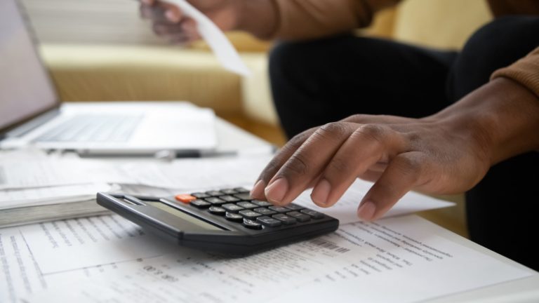 Afro Caribbean man sitting on modern yellow sofa at low table with paperwork and laptop cropped to show hand pressing buttons on calculator