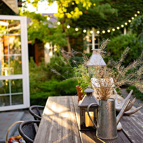 Pretty summer courtyard garden with open patio doors and string lights wooden table and wicker chairs with dried sprig arrangement in a metal ornate watering can and selection of outdoor lanterns