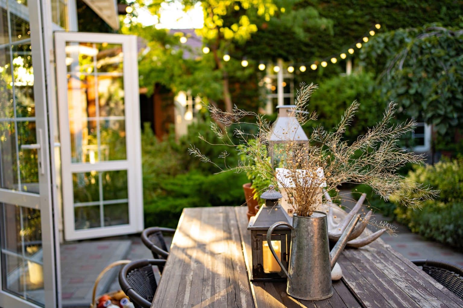 Pretty summer courtyard garden with open patio doors and string lights wooden table and wicker chairs with dried sprig arrangement in a metal ornate watering can and selection of outdoor lanterns