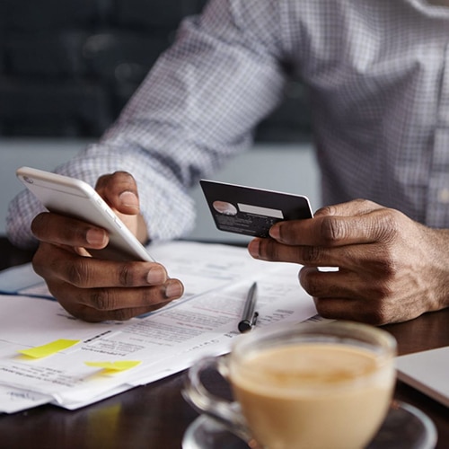 Afro Caribbean man sat at dark wood desk with laptop paperwork pen and coffee holding bank card in one hand and mobile phone in the other