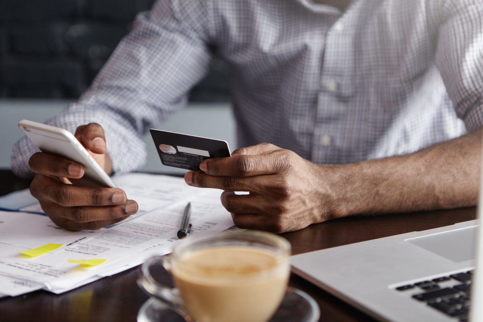 Afro Caribbean man sat at dark wood desk with laptop paperwork pen and coffee holding bank card in one hand and mobile phone in the other