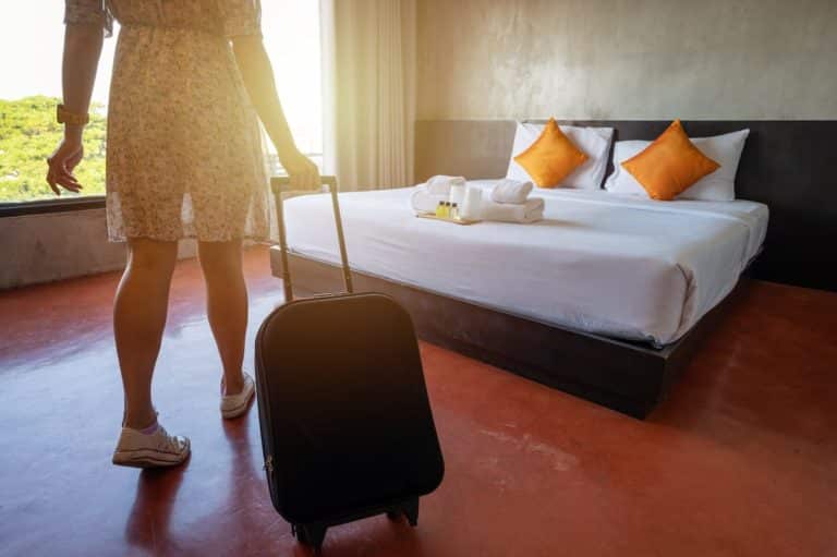 King size hotel bedroom with towels and pamper tray on white bedding with woman in summer dress and trainers with small suitcase on wheels