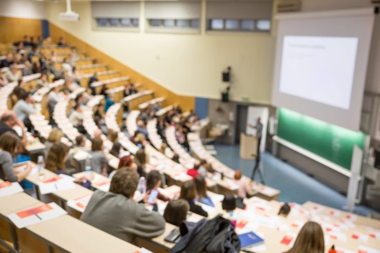 Students in auditorium with lecturer at lecturn in front of large projector screen